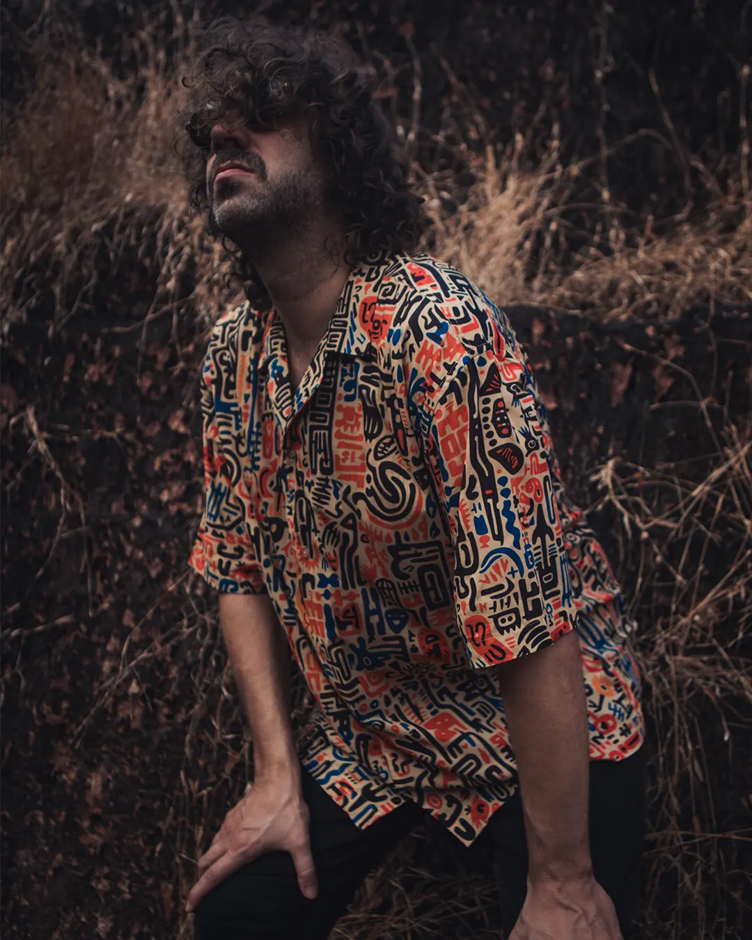 Person wearing a colorful patterned shirt sitting in a natural setting with dry grass.