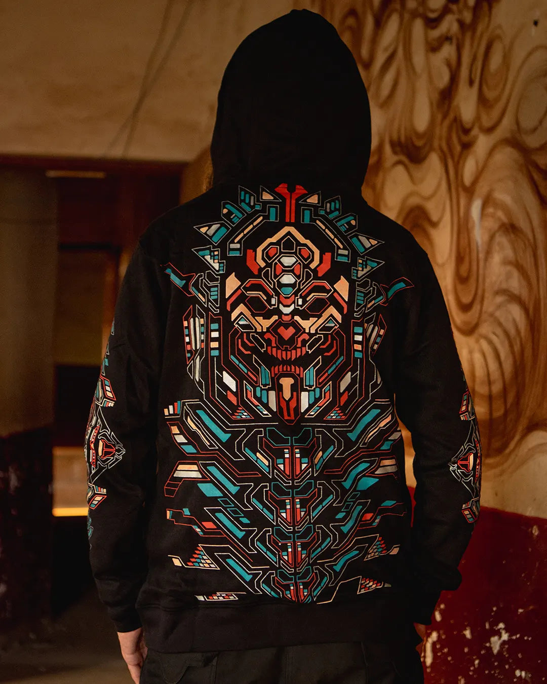 Person wearing a black hoodie with a colorful geometric design on the back, standing against a textured wall.