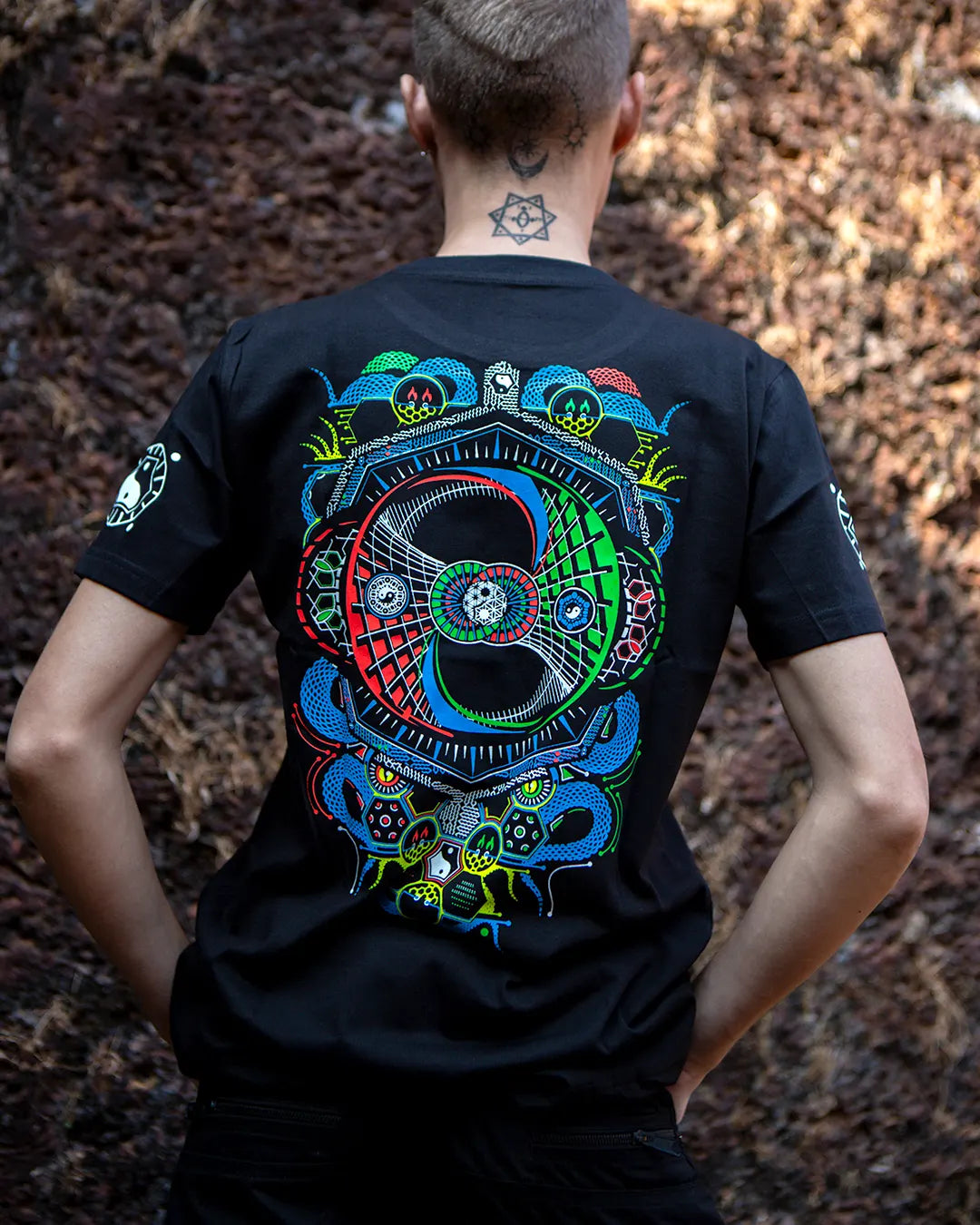 Person wearing a black t-shirt of ultra tribe brand with a colorful abstract ying yang Damaru design with glow in dark prints on the back, standing against a natural background.