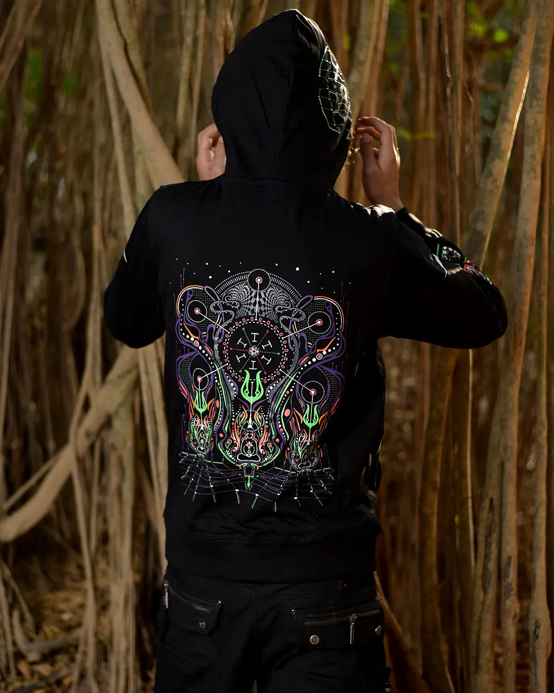 A person wearing a ultra tribe clothing spiritual network uv light reactive black hooded sweatshirt with a large colorful psychedelic graphic on the back, standing in front of a dense background of hanging vines.