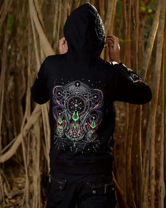 A person wearing a ultra tribe clothing spiritual network uv light reactive black hooded sweatshirt with a large colorful psychedelic graphic on the back, standing in front of a dense background of hanging vines.
