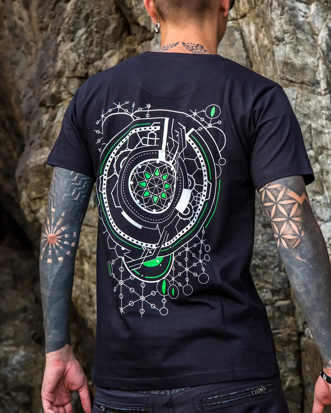 Person wearing a black t-shirt with a detailed design on the back, standing against a stone wall.