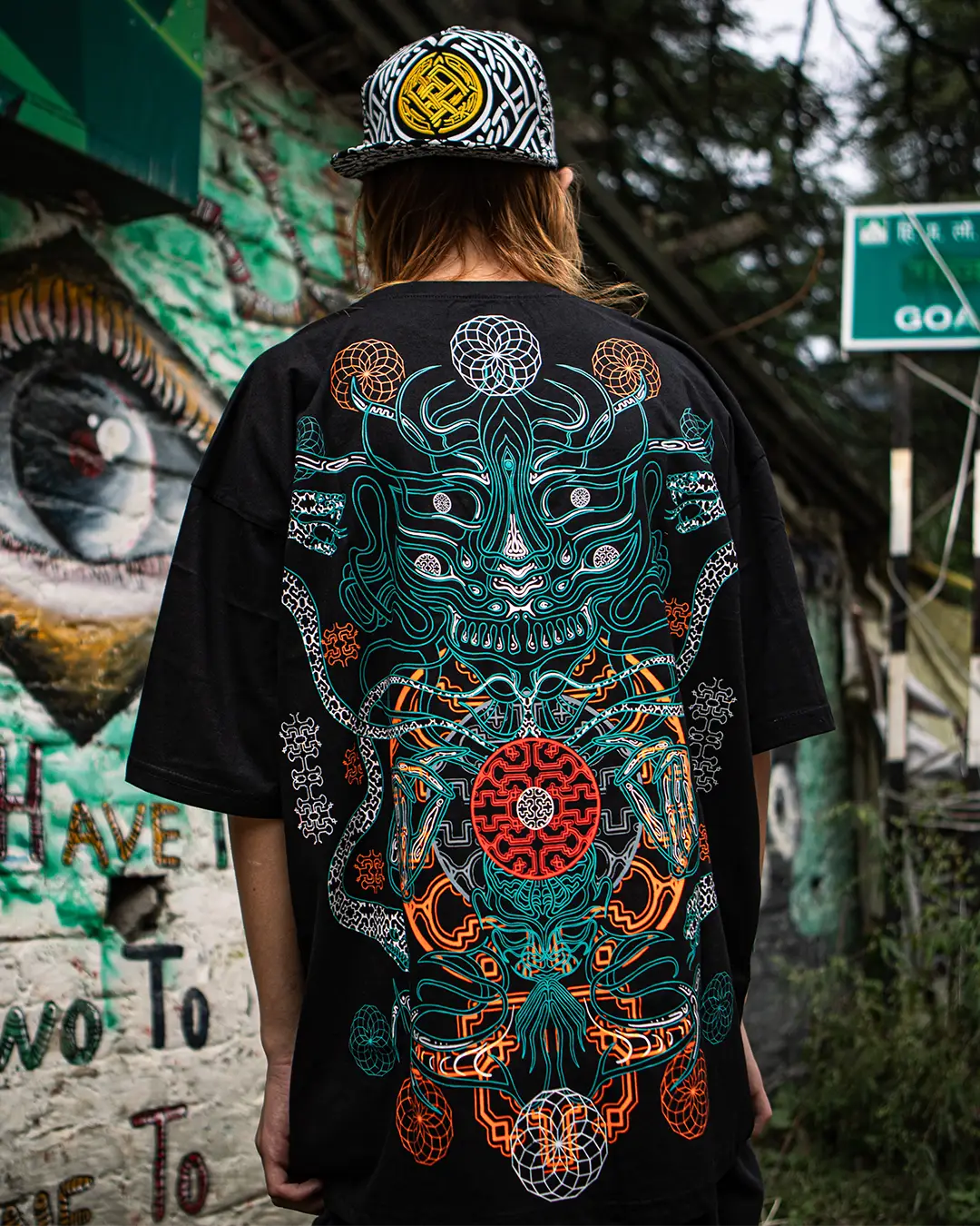 Person wearing a black t-shirt with a colorful design on the back, standing in front of graffiti.