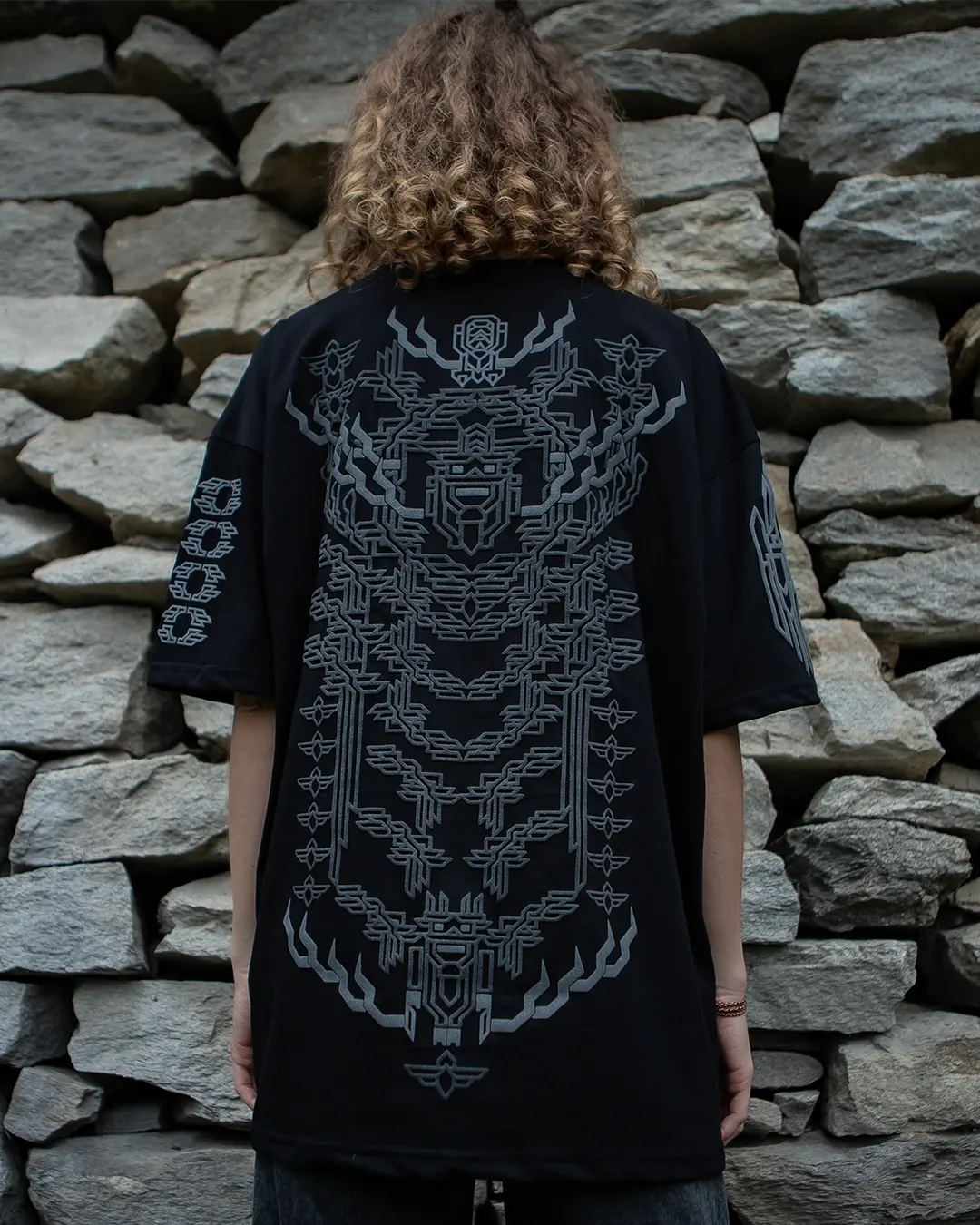 Person wearing a black t-shirt with intricate Protector 3dpuff design by ultratribe white design against a stone wall.