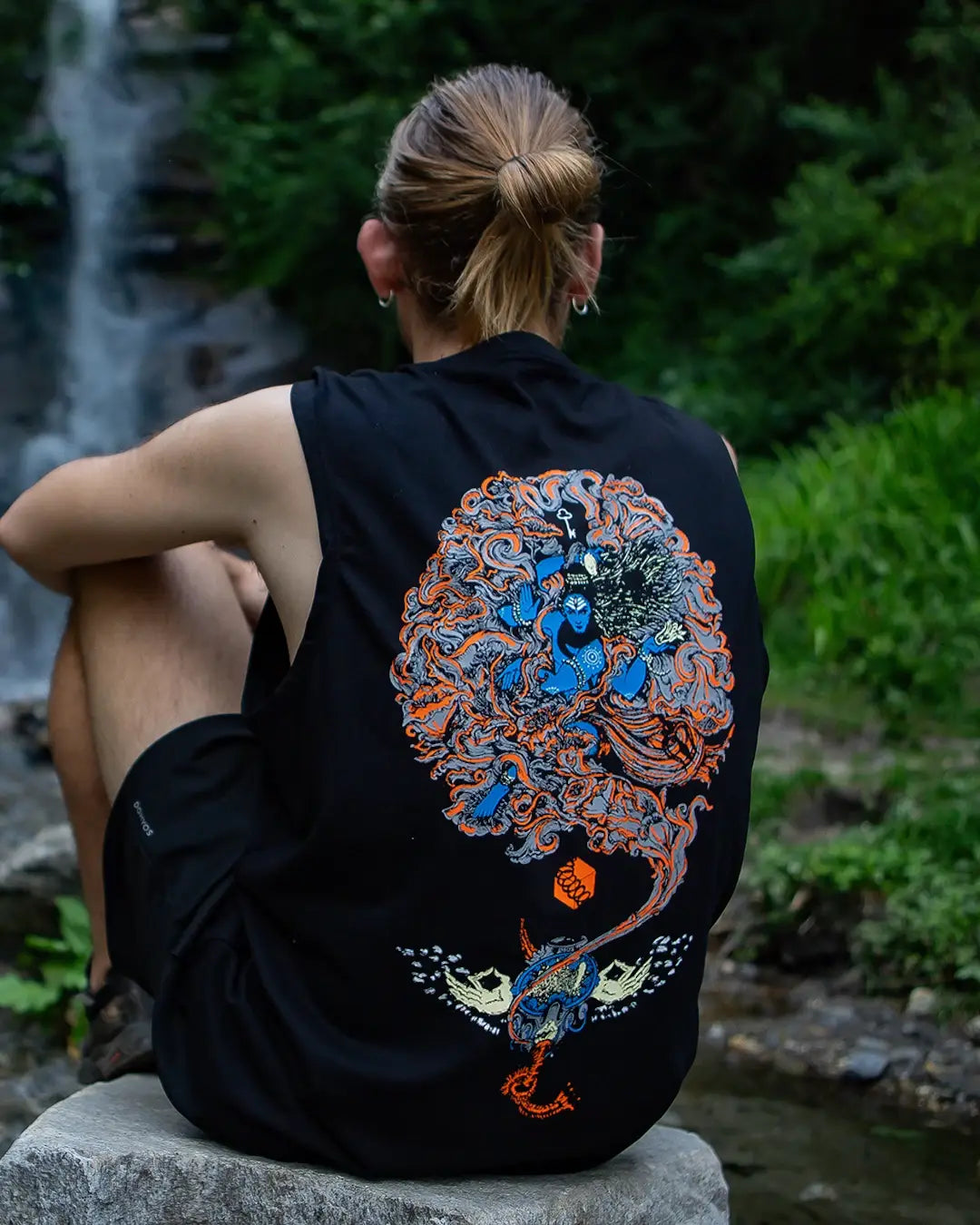 Person wearing a black sleeveless shirt with a colorful dragon design sitting by a waterfall.
