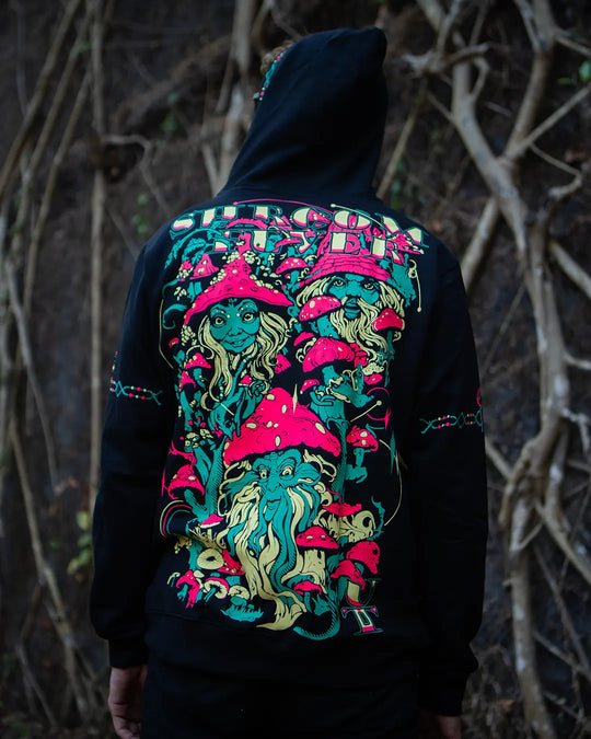 Person wearing a black hoodie with a colorful mushroom and gnome design on the back, standing in a natural setting.
