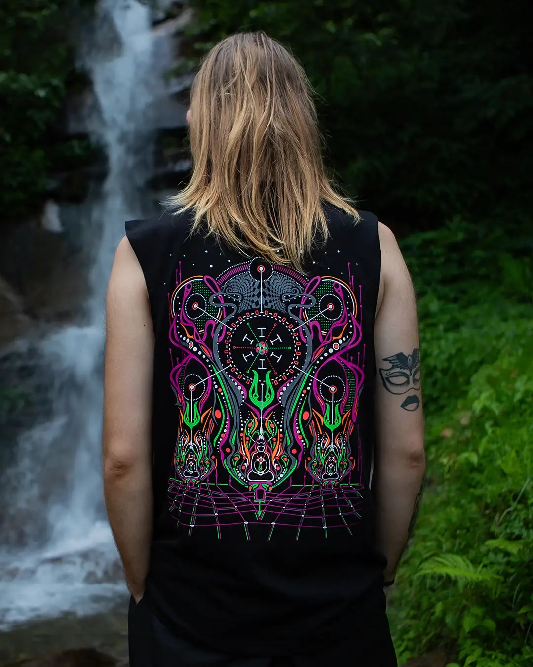 Person wearing a black sleeveless shirt with a colorful abstract design, standing in front of a waterfall.