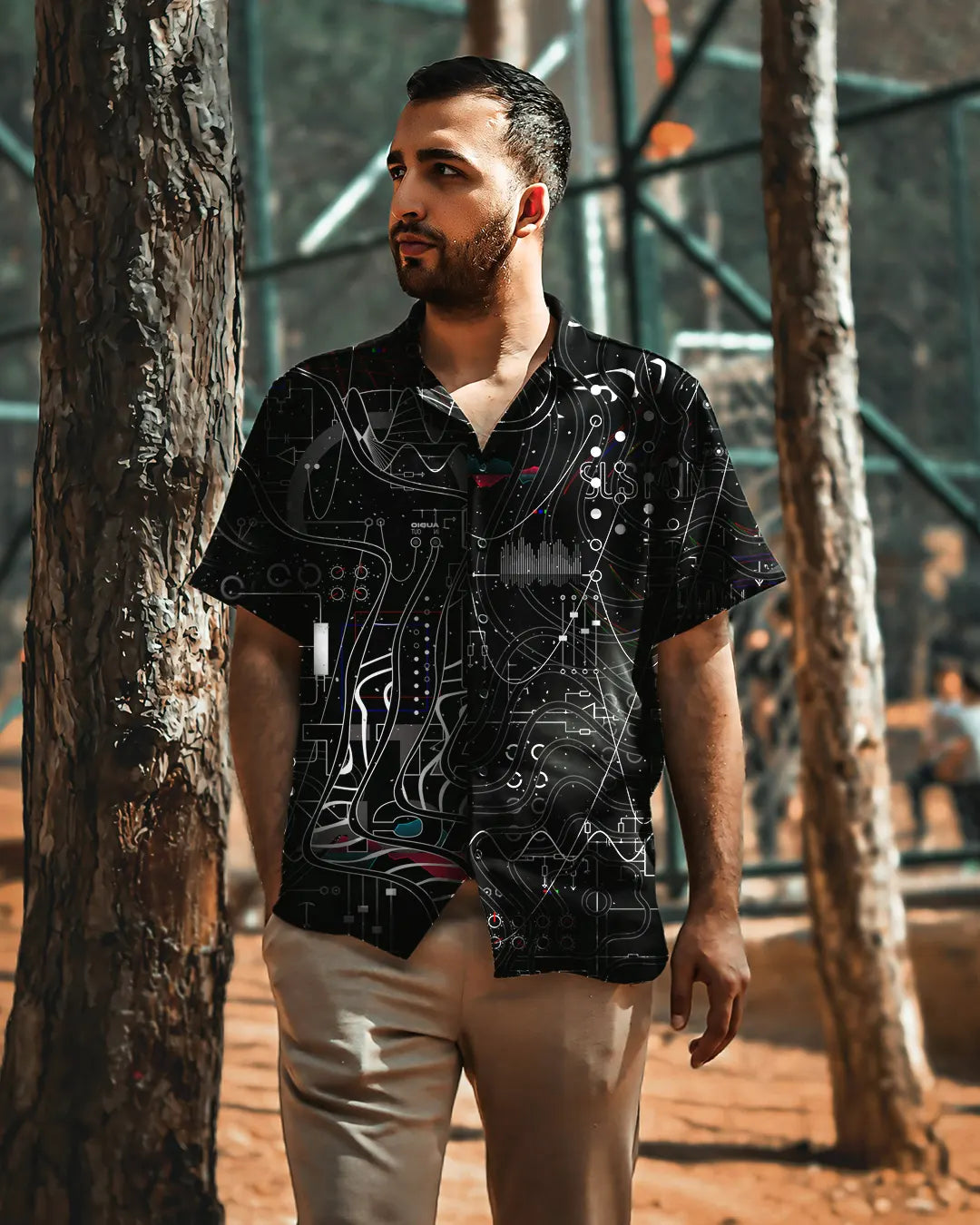 Man wearing a black patterned shirt standing between two trees.