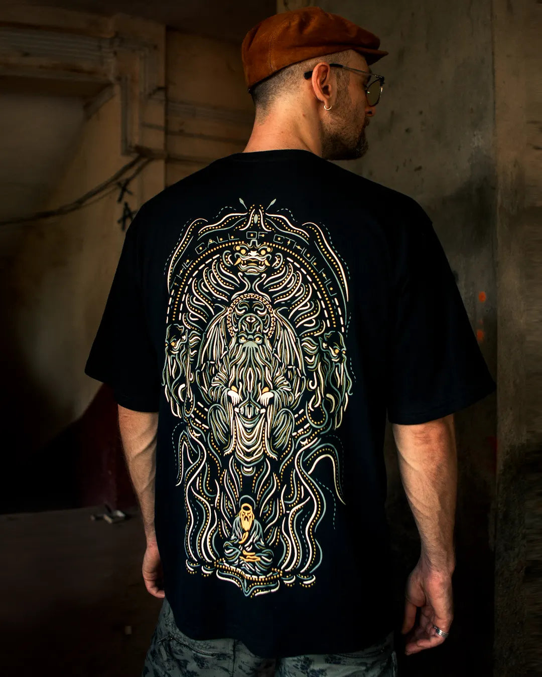 Man wearing a ultra tribe gow in dark and uv light reactive black oversized t-shirt with a detailed psuchedelic design the call of cthulhu design on the back, standing in a dimly lit room.