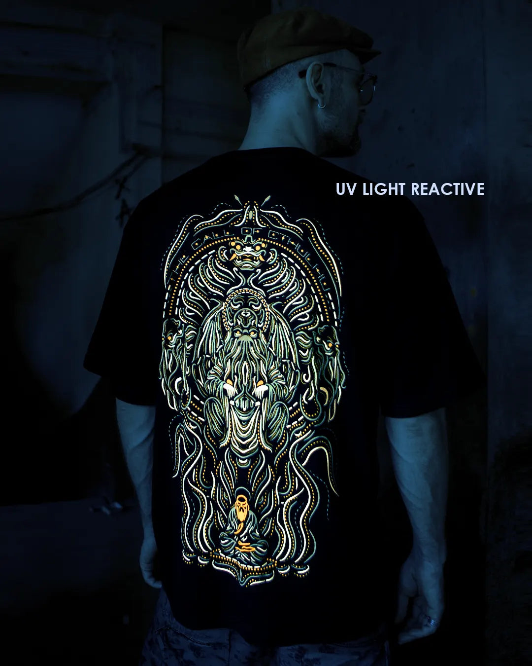 Person wearing a ultra tribe gow in dark and uv light reactive black oversized t-shirt with a detailed psuchedelic design the call of cthulhu design black t-shirt with a UV light reactive design, standing in a dark setting.