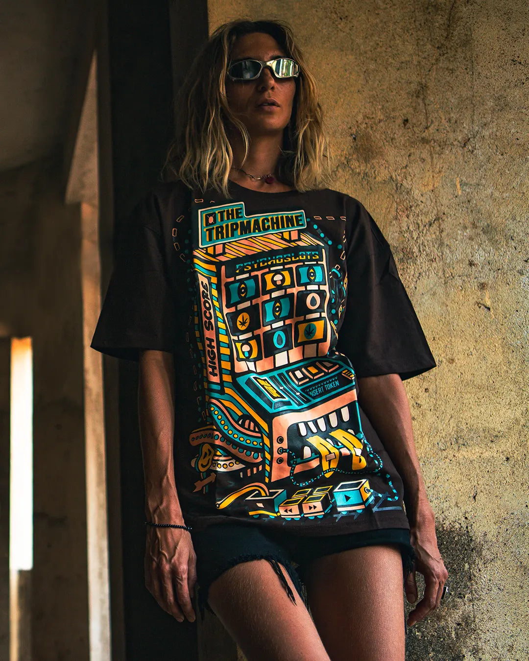 a women wearing a ultra tribe brand dark brown t-shirt with a colorful psychedleic and UV Light Reactive graphic design against a textured wall.