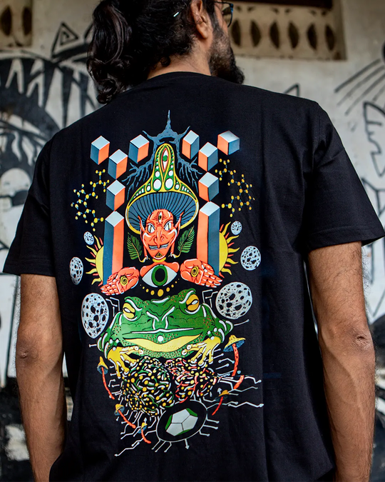 a Person wearing a black ultra tribe uv light reactive plus glow in dark t-shirt with a colorful triptamind graphic design on the back, standing against a graffiti-covered wall.