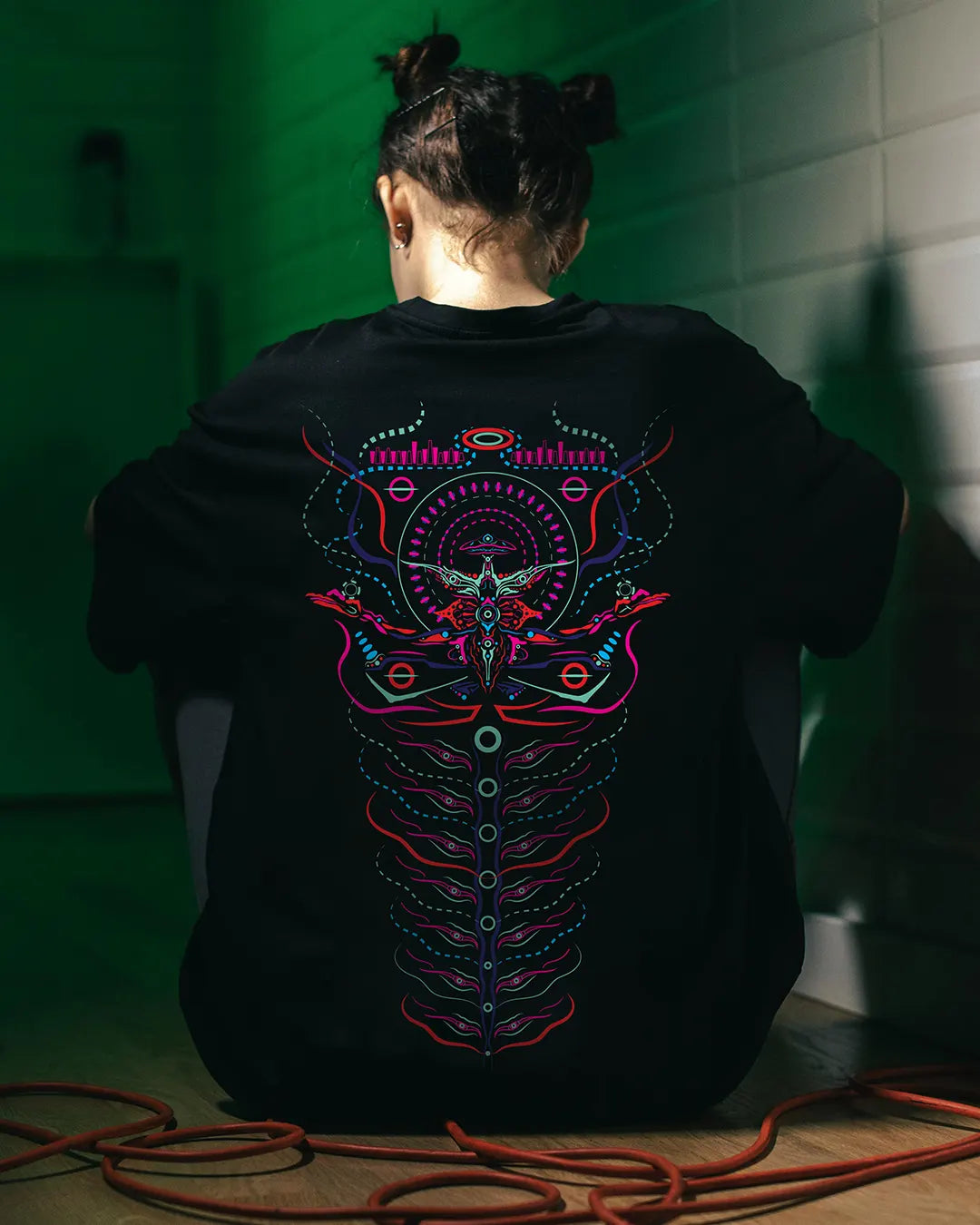 a women wearing a black t-shirt with colorful abstract design on the back, sitting against a green wall. design by ultra tribe uv light reactive prints tshirt 