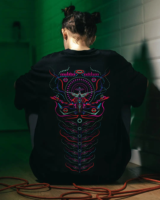 a women wearing a black t-shirt with colorful abstract design on the back, sitting against a green wall. design by ultra tribe uv light reactive prints tshirt 
